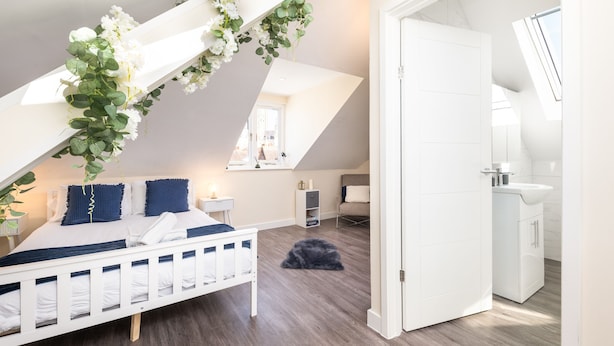 Une chambre avec un lit blanc, une petite table de chevet et une commode. Il y a une fenêtre qui donne sur l’extérieur.