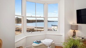 Una ventana saliente con vista a una playa y al mar, un pequeño banco con libros, y una lámpara sobre una mesita auxiliar.