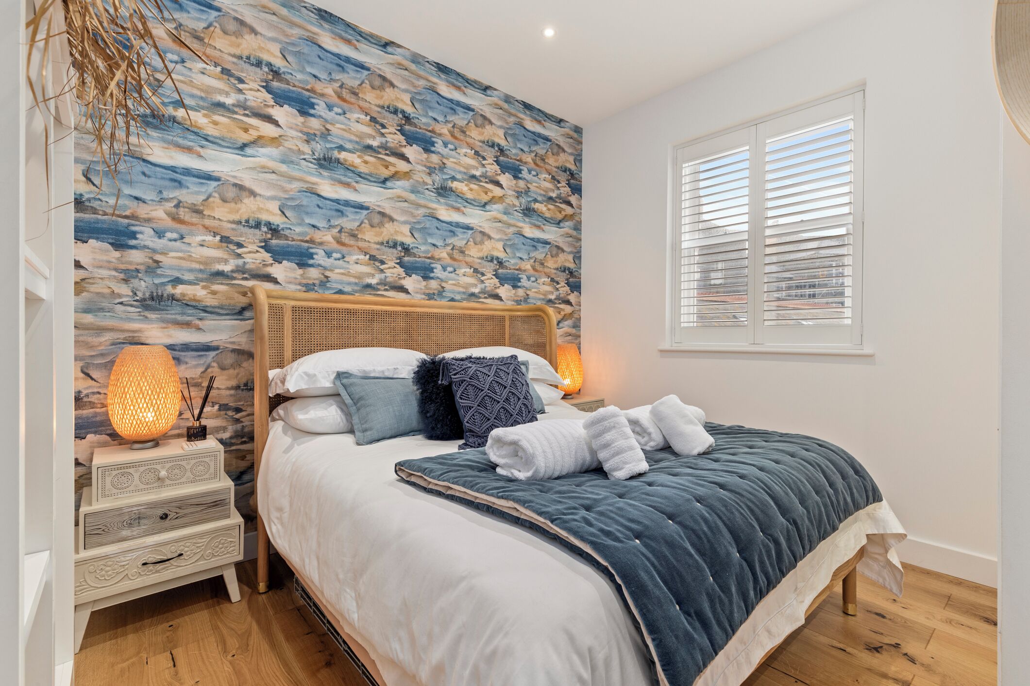 A bedroom with a bed, a nightstand, a wall-mounted lamp, and a window with blinds.