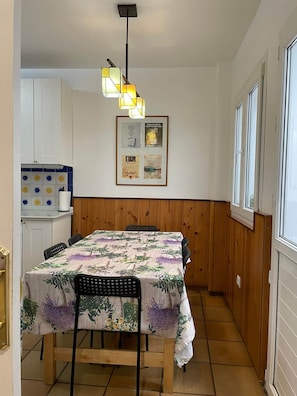 A dining area with a table covered in a floral tablecloth, wooden chairs, and a hanging light fixture.