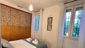 A bedroom with a bed, a bench, two windows with curtains, and a wall with framed pictures. - Plaza España Family House, amplia casa con parking gratuito en la calle, ideal para familias y grupo... (Seville)