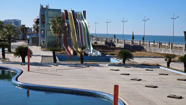 A swimming pool with a water slide, palm trees, and a beach in the background. - Kale Park  (Batumi)