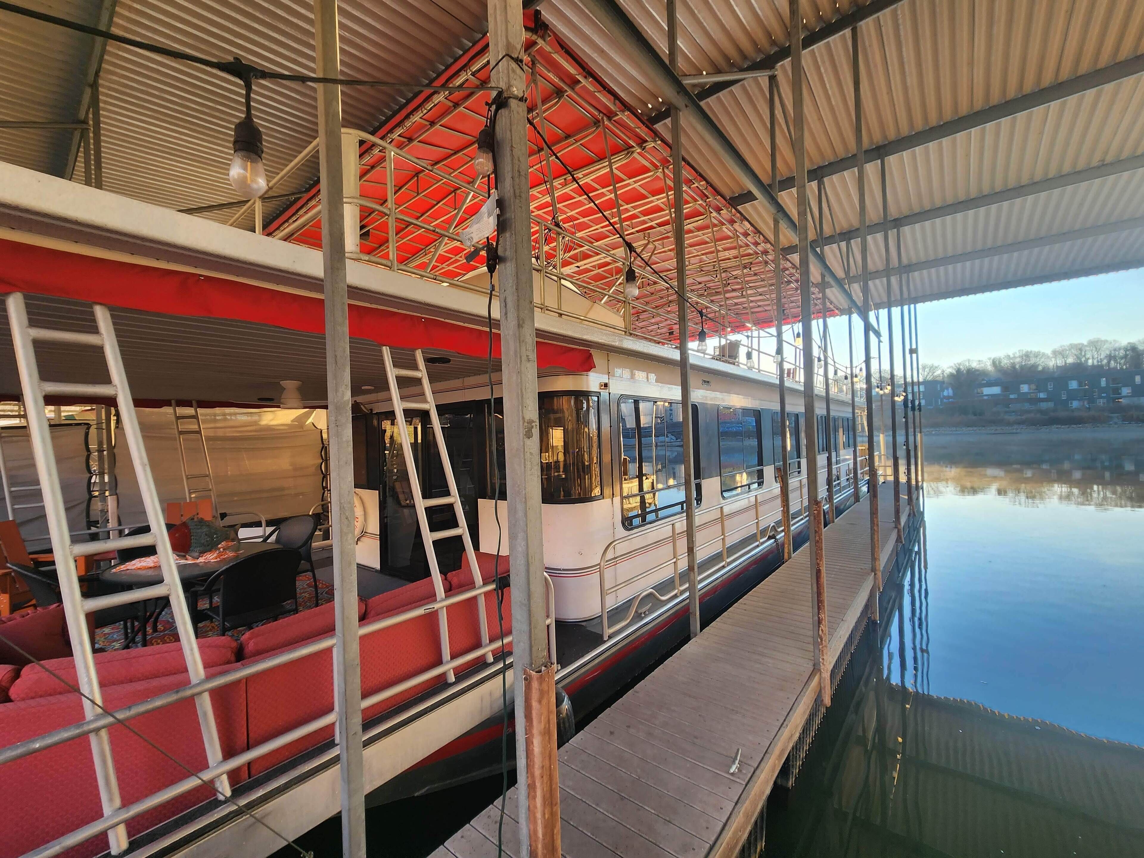 A river cruise ship with a covered upper deck, red seating, and a wooden deck.