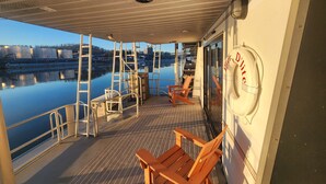 A covered deck with wooden chairs and tables, a lifebuoy, and a view of a calm river with buildings in the background. - Pier D Lite 4 bdrm Houseboat on River Downtown Knoxville - walk to Univ. of TN  (Knoxville)