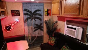 Een kamer met een rode neon-gitaar, een zwarte bank met kleurrijke kussens, een houten kast en een grote plant.