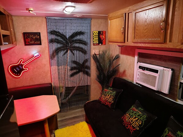 Um quarto com um letreiro de guitarra de neon vermelho, um sofá preto com almofadas coloridas, um armário de madeira e uma planta grande.