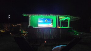 Área de bar ao ar livre com uma grande tela exibindo uma cena de praia, decorada com luzes de corda e mobiliário de madeira.