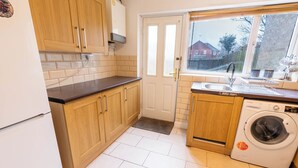 Cocina compacta con armarios de madera, lavadora integrada y ventana con vistas a una casa.