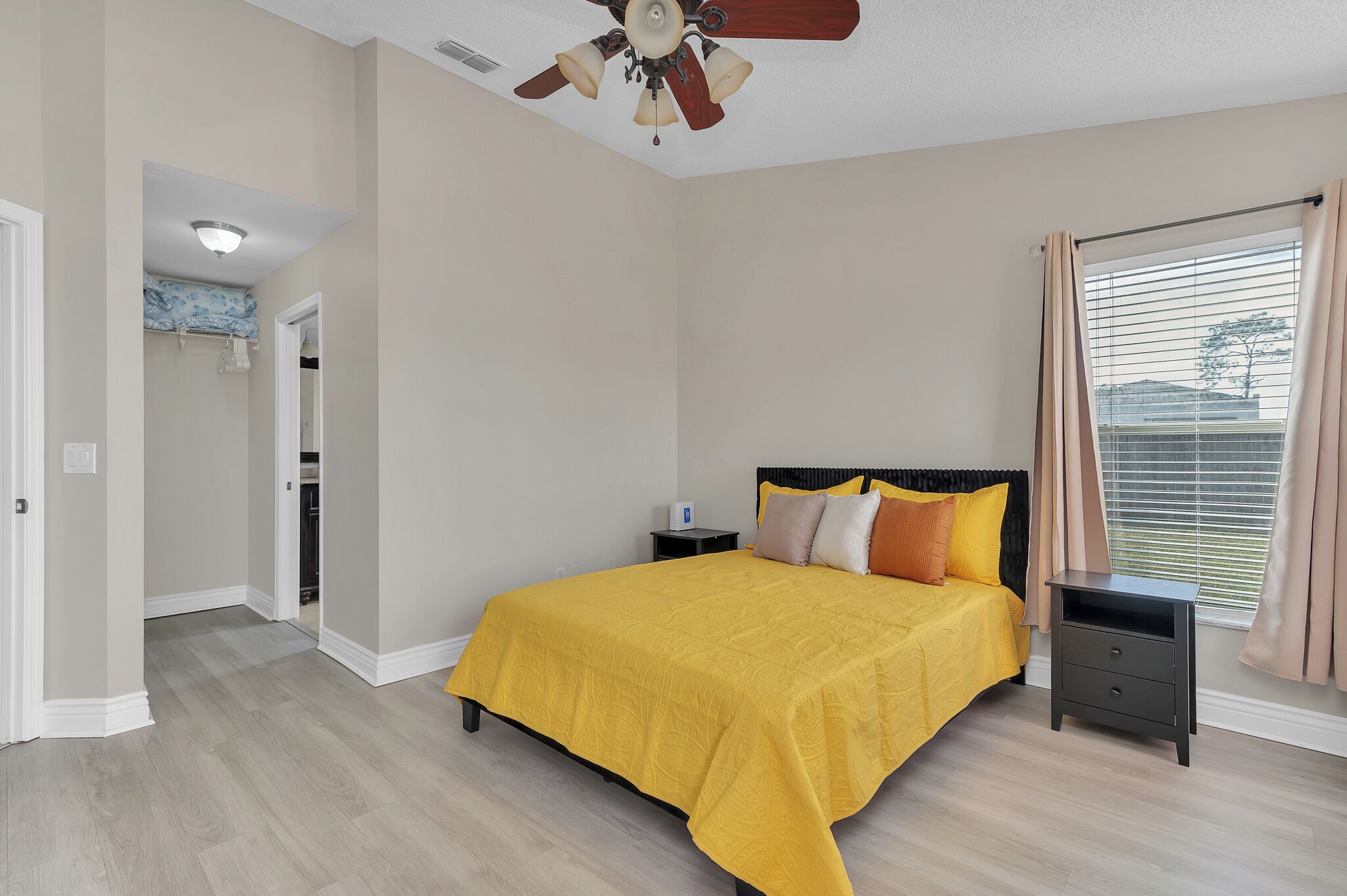 Une chambre à coucher comprenant un lit, une table de chevet, un ventilateur de plafond, une fenêtre avec des rideaux et une porte donnant sur une autre pièce.