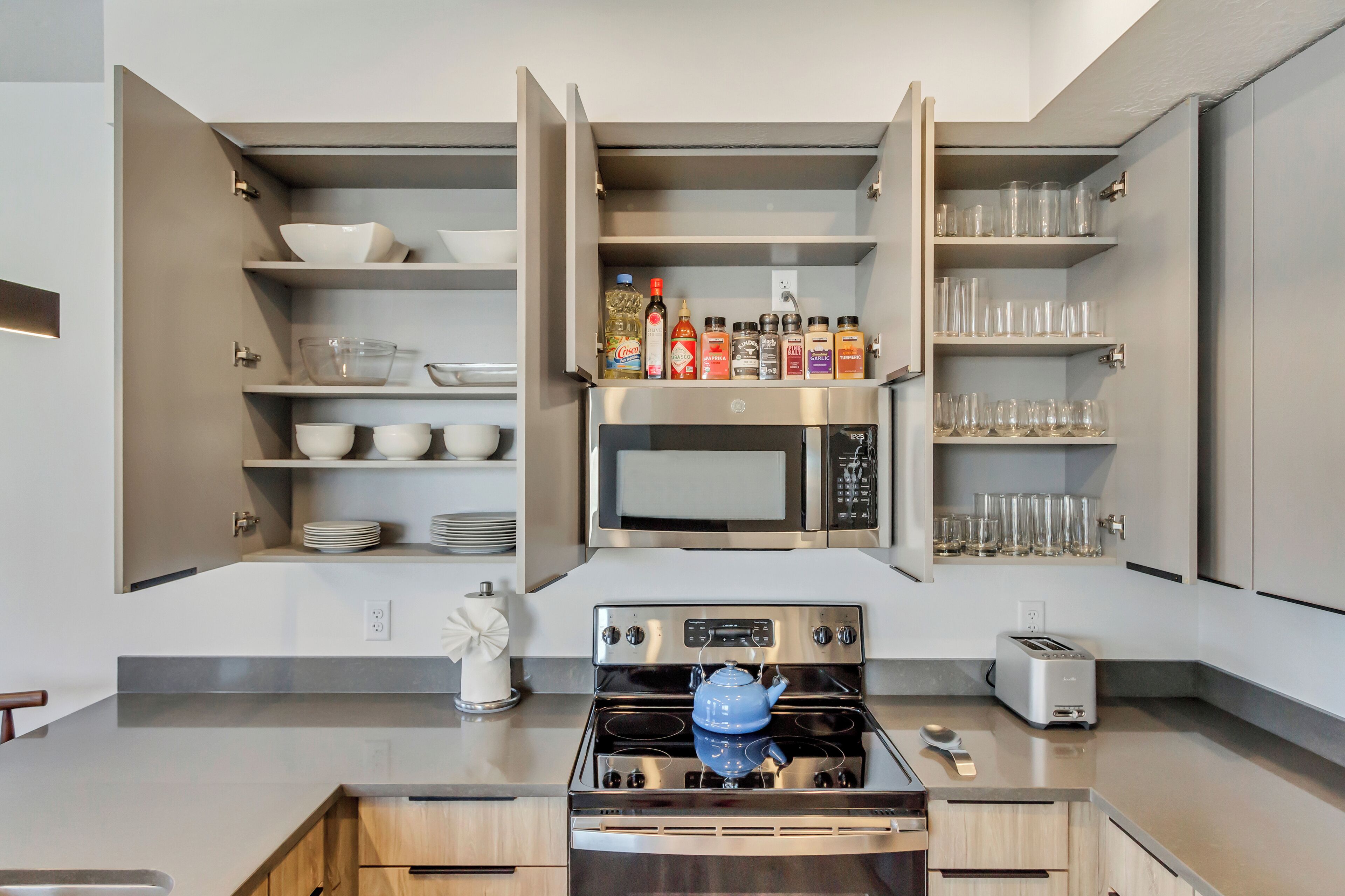 Une cuisine moderne dotée d’appareils électroménagers intégrés, d’étagères ouvertes où sont exposées la vaisselle et les verres, et d’une bouilloire bleue posée sur la cuisinière.