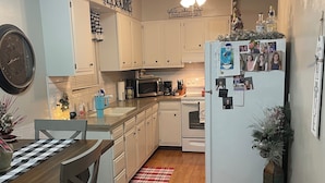 A compact kitchen with white cabinets, a microwave, a stove, and a refrigerator adorned with photos. A dining table with chairs is positioned in the foreground. - Charming 2-bedroom house in delightful Jackson with AC, WiFi (Jackson)