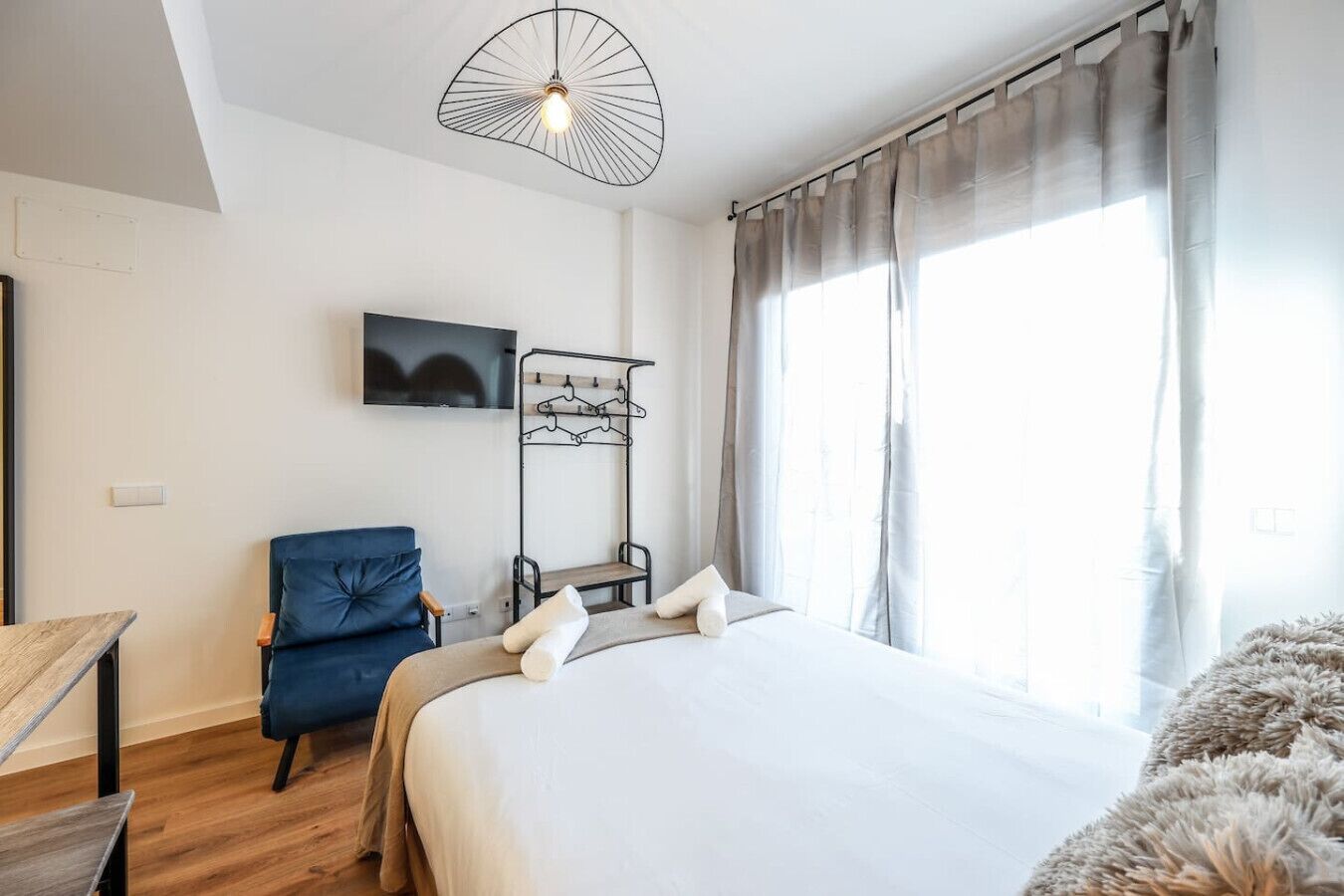 Un dormitorio con cama, una silla, un televisor, perchero y ventana con cortinas.