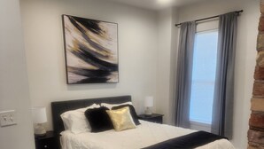 Une chambre à coucher avec un lit, une table de chevet, une lampe, une fenêtre avec des rideaux et un tableau au mur.