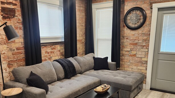 Un salon moderne avec un canapé d’angle gris, une table basse noire et un mur en briques.