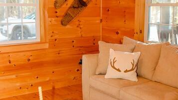 Un interior de cabaña acogedor, con piso de madera, un sofá con un cojín con forma de ciervo y un par de botas colgadas en la pared.