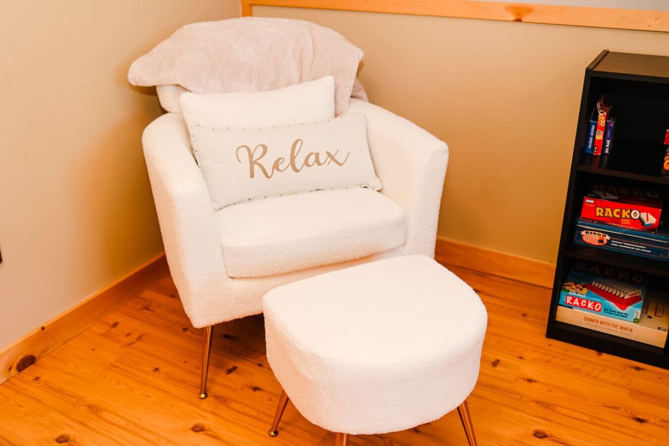 A white chair with a 'Relax' pillow and a matching ottoman, positioned next to a bookshelf with board games.