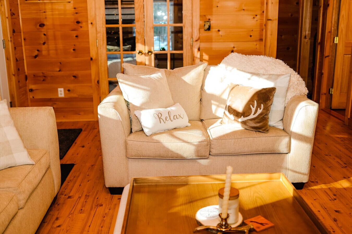 Interior acogedor de una cabaña con un sofá color beige, una mesita con una vela y una puerta de madera al fondo.