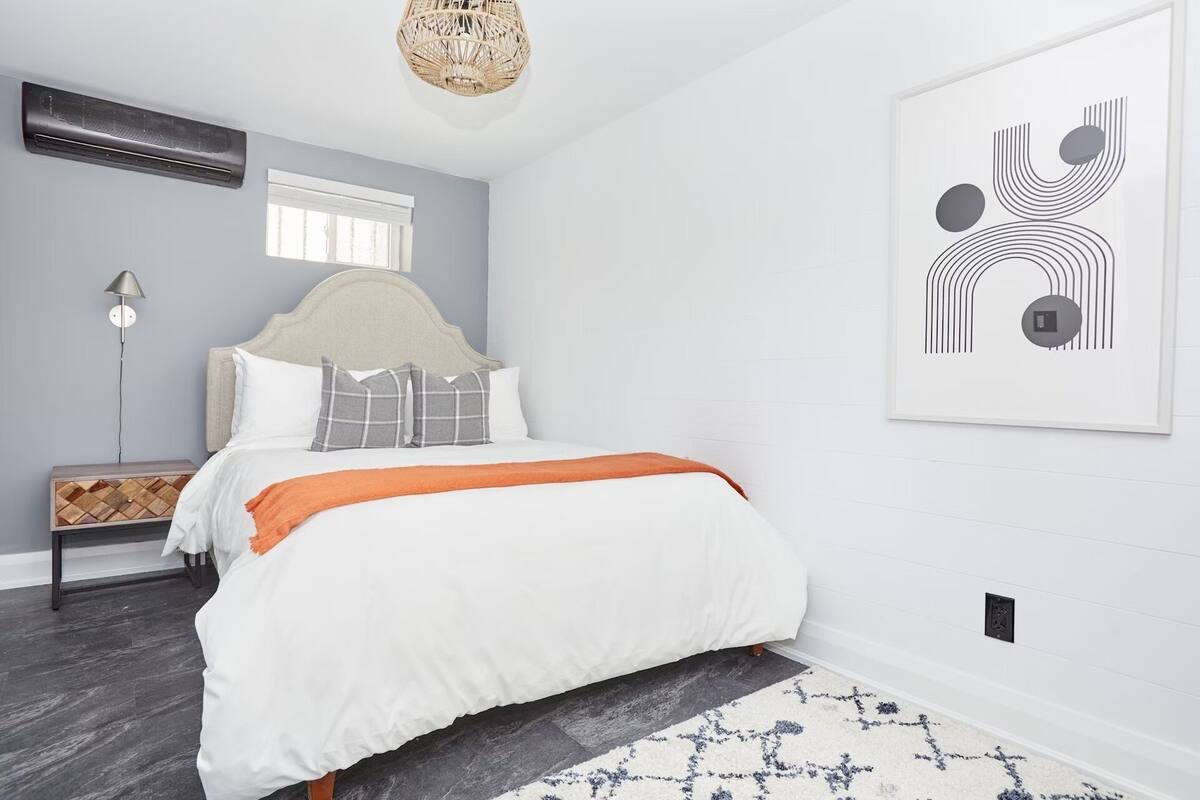 A bedroom with a bed, a bedside table, a lamp, a wall-mounted air conditioner, a hanging light fixture, and a framed abstract artwork.