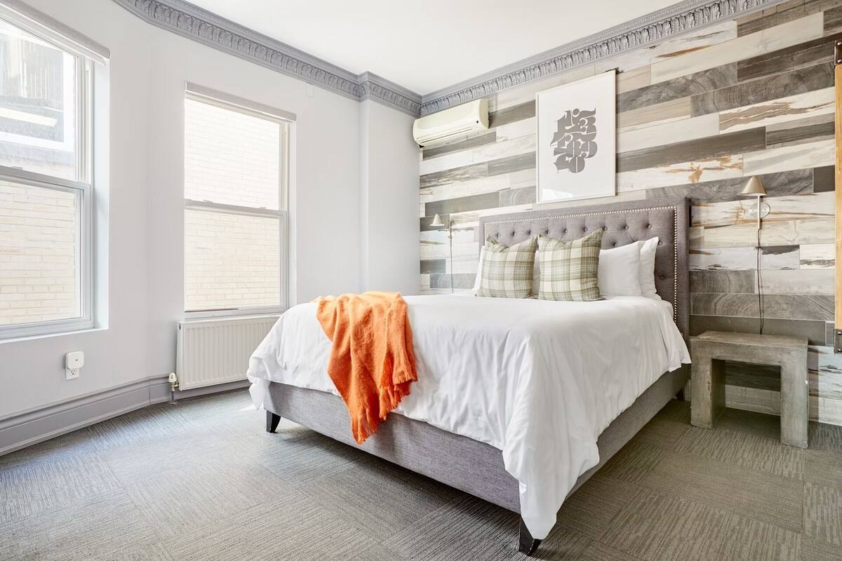 A bedroom with a bed, a nightstand, a wall-mounted air conditioner, and a window with blinds.