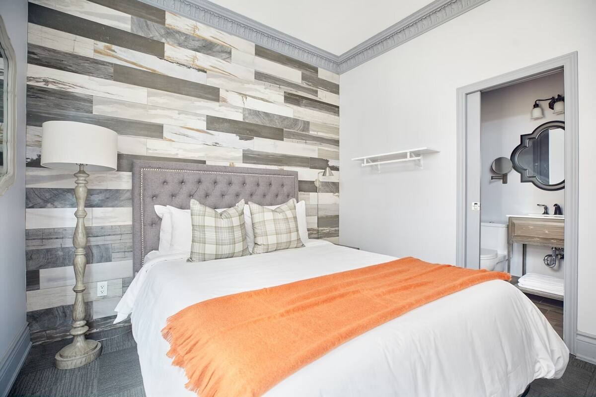 A hotel room with a bed, a nightstand, a lamp, a mirror, and a bathroom visible through an open door.