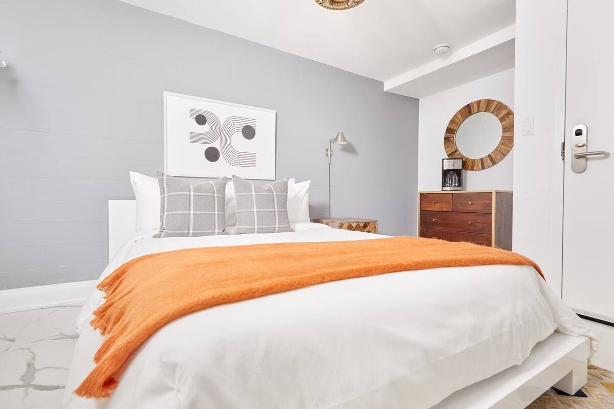 A modern bedroom with a bed, a bedside table, a lamp, a dresser, and a door.
