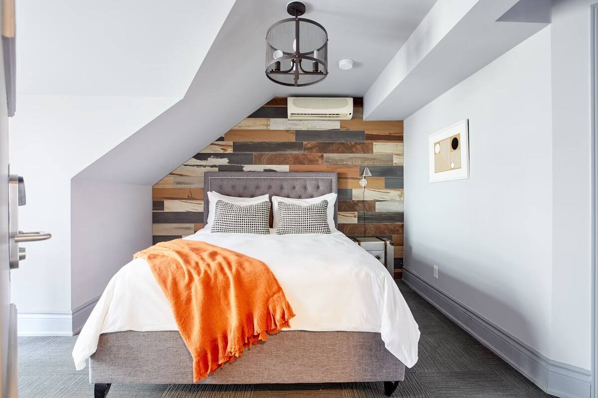 A bedroom with a sloped ceiling, a bed with a grey headboard, a white and orange throw blanket, and a modern pendant light.