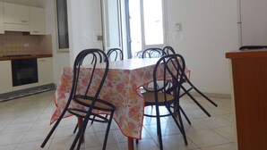 Un comedor con una mesa cubierta por un mantel floral, rodeada por seis sillas. Se ve a la izquierda una zona de cocina con armarios y una estufa.