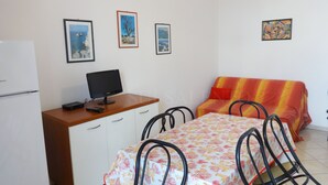 Una habitación pequeña y bien iluminada con una cama, un escritorio con computadora, una silla y una mesa de comedor. Hay tres cuadros en la pared.