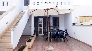 Un edificio de dos pisos con una escalera, un área de estar al aire libre techada con sillas y una sombrilla, y una mesa pequeña.