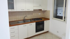 Una pequeña cocina con armarios blancos, cocina a gas, un lavabo y una ventana con persianas.