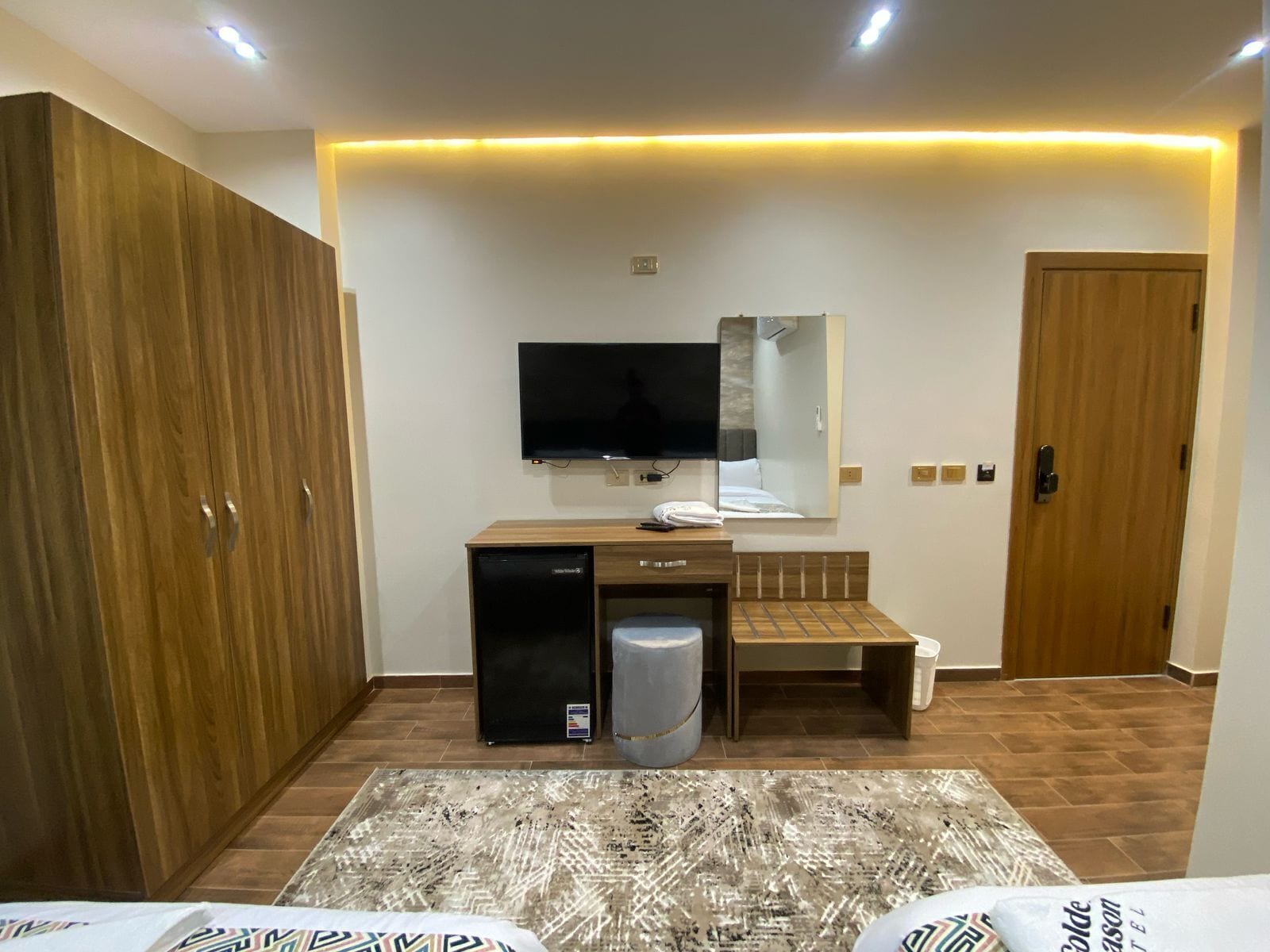 Habitación de hotel moderna con un armario de madera, un escritorio pequeño con refrigerador, televisor de pantalla plana montado en la pared y una mesita de noche con espejo.
