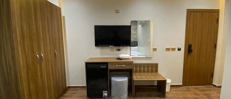 Habitación de hotel moderna con un armario de madera, un escritorio pequeño con refrigerador, televisor de pantalla plana montado en la pared y una mesita de noche con espejo.