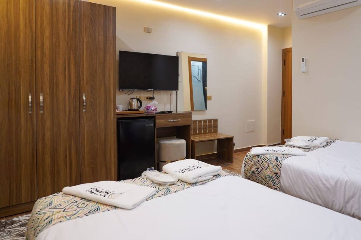 A hotel room with two beds, a wardrobe, a desk with a chair, a television, and a small kitchenette.