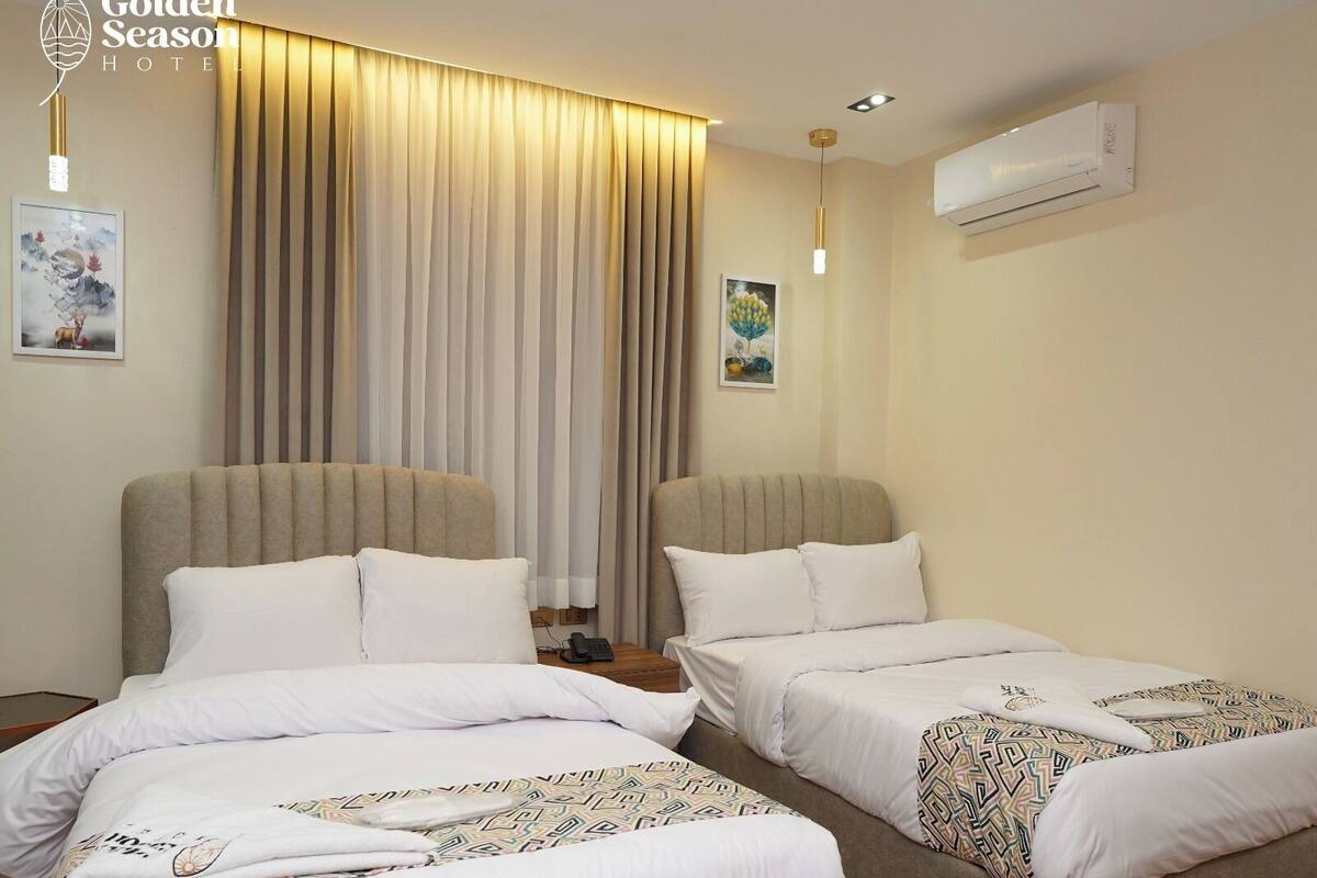 A hotel room with two beds, beige curtains, a painting, and an air conditioning unit.