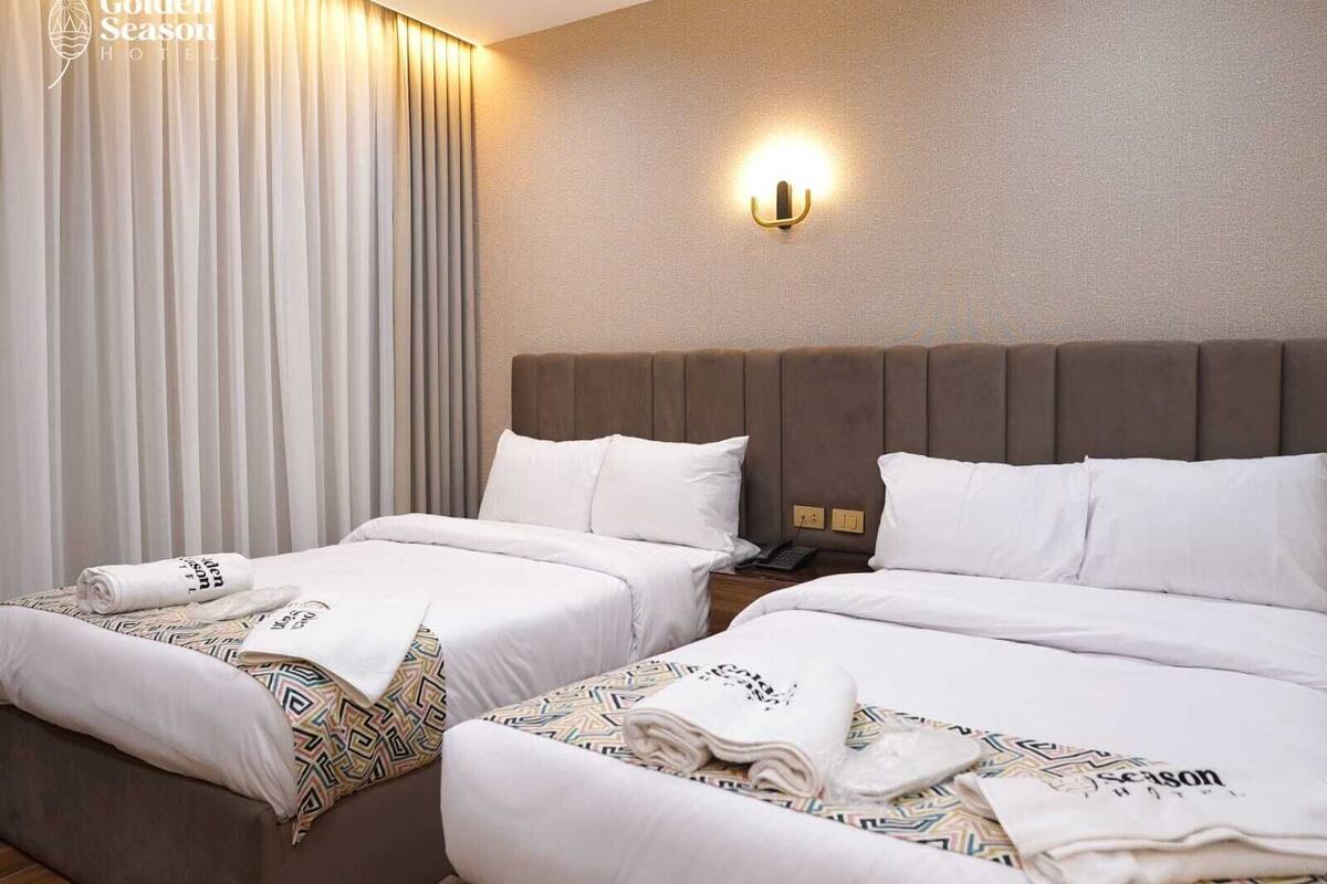 A hotel room with two beds, a headboard, a nightstand, a lamp, and a window with curtains.