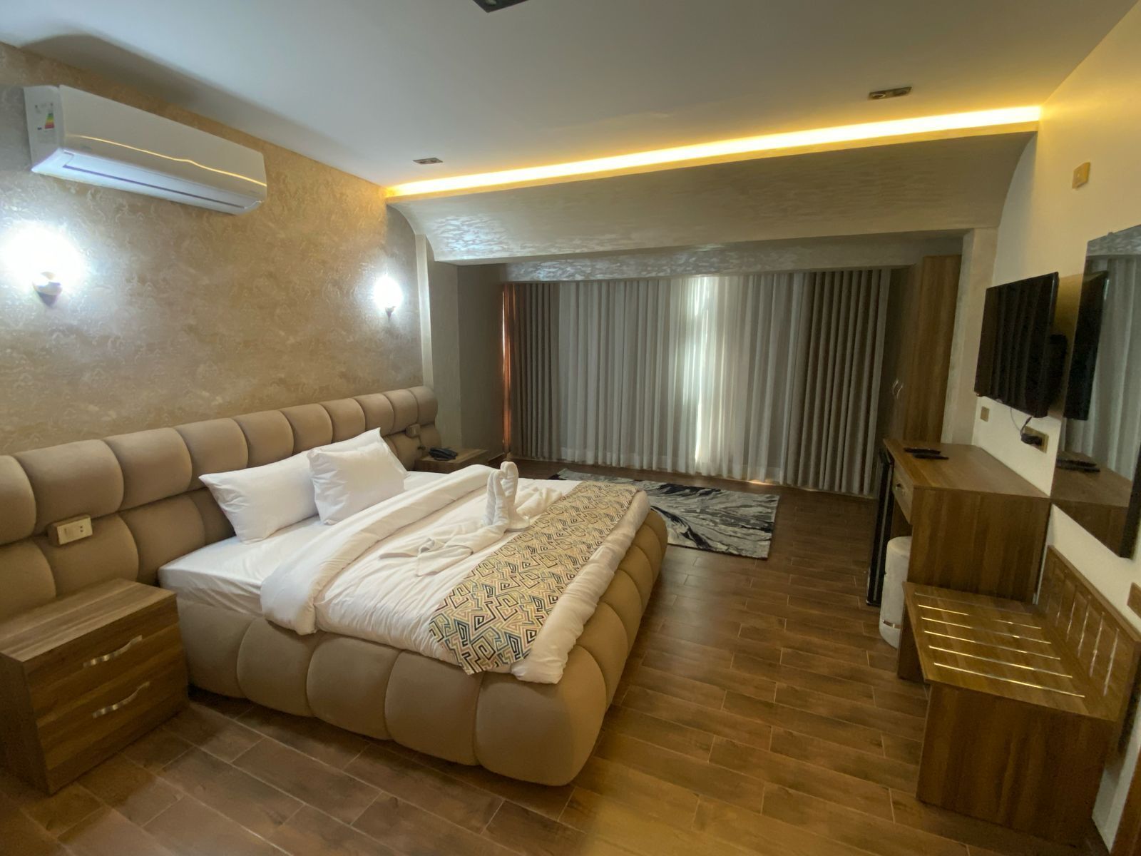 Una habitación de hotel moderna con una cama grande, un tocador de madera, un televisor y un aire acondicionado instalado en el techo.