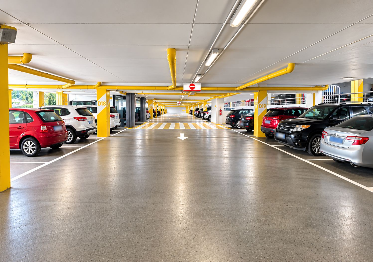 Un parking à étages avec plusieurs rangées de voitures garées, des piliers de soutien jaunes et des flèches directionnelles peintes sur le sol.