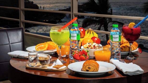 Una mesa dispuesta con alimentos para el desayuno, incluyendo frutas, jugos y un croissant, con vista a un atardecer.