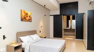 Quarto de hotel moderno com cama, mesas de cabeceira, uma pintura, um guarda-roupa e um banheiro visível por uma porta aberta.