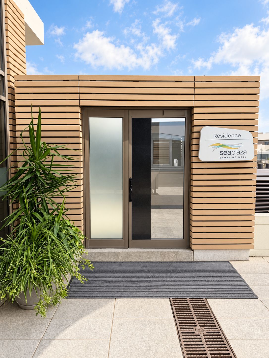 Une entrée moderne avec une porte en verre, une enseigne indiquant « Résidence Seaplaza, centre commercial », et un pot de fleurs placé à côté.