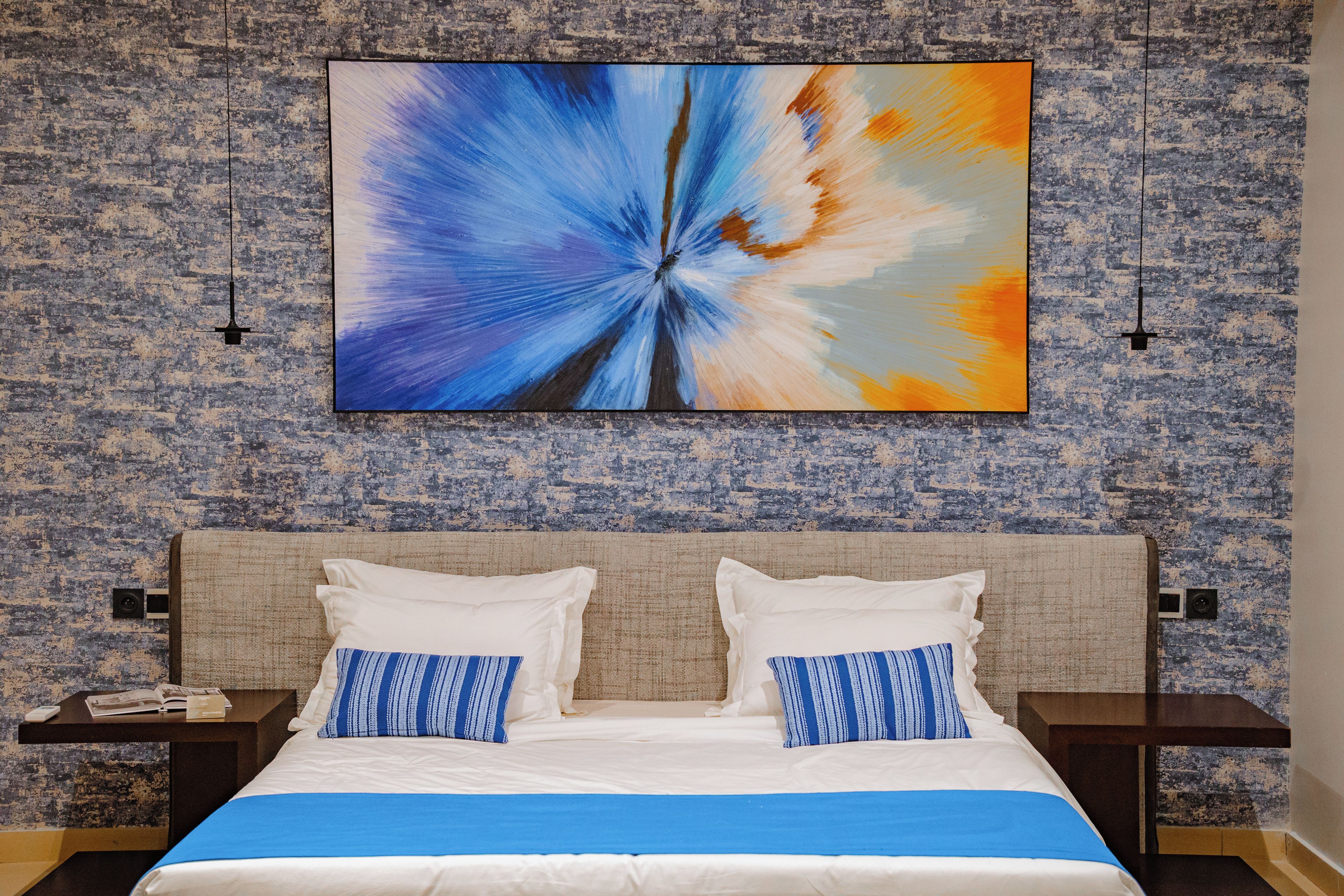 A hotel room with a large abstract painting, a bed with white and blue bedding, and two nightstands with lamps.