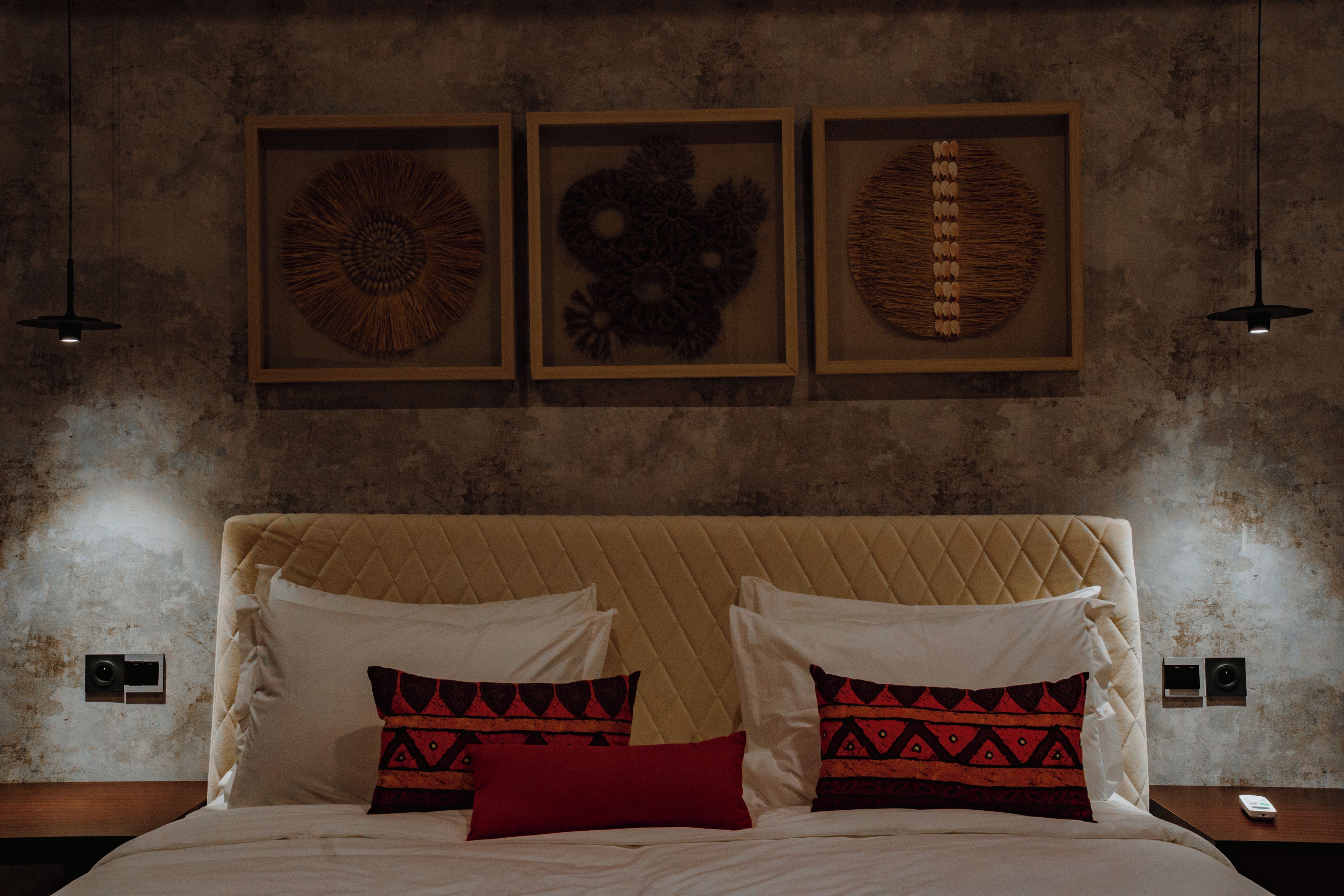 Cama com cabeceira acolchoada, dois travesseiros brancos e dois travesseiros estampados. Acima da cama, três obras de arte emolduradas na parede.