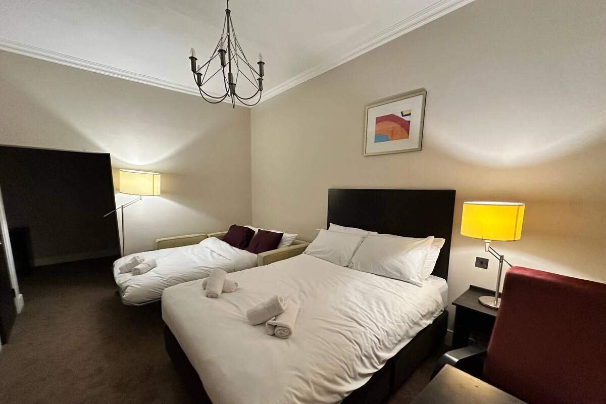 Ein Hotelzimmer mit zwei Betten, einer Kandelaber, einem Gemälde und einem roten Sessel.