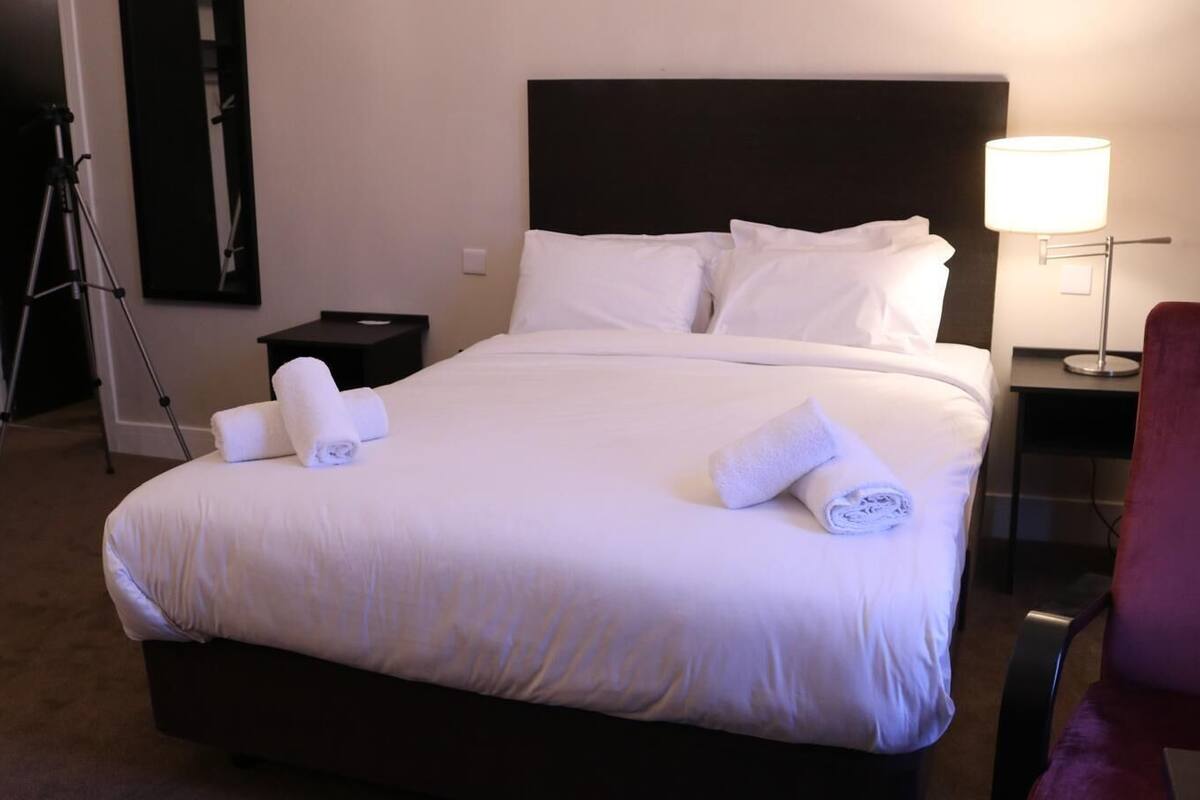 Ein Hotelzimmer mit einem großen Bett, zwei Nachttischen, einer Lampe, einem Sessel und einem Stativ.