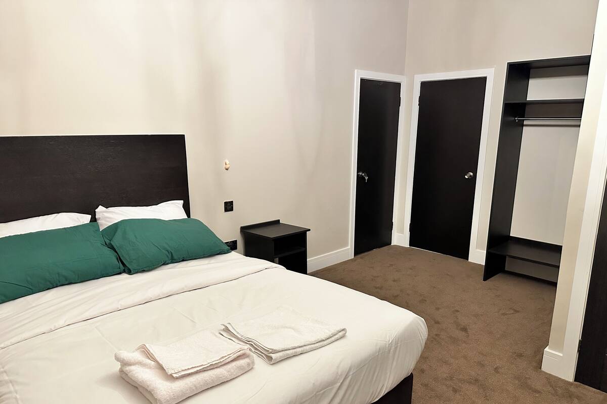 Ein ordentlich bezogenes Bett mit weißen und grünen Kissen, ein kleiner Nachttisch und ein Satz Handtücher. Zwei schwarze Türen und ein Schrank mit Regalen sind ebenfalls zu sehen.