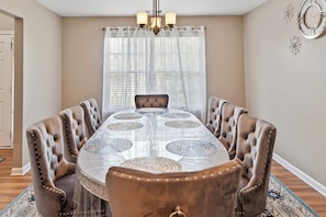 A dining room with a long table, upholstered chairs, a chandelier, and a window with sheer curtains.