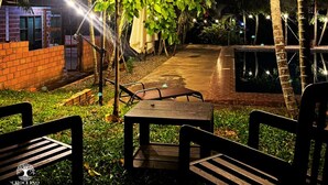 An outdoor seating area with tables and benches, surrounded by greenery and palm trees, under string lights.