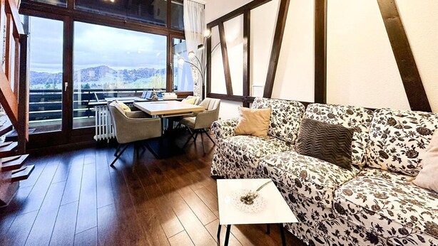 Ein modernes Wohnzimmer mit Holzboden, einem floralen Sofa, einem Esstisch mit Stühlen und großen Fenstern mit Blick ins Freie.