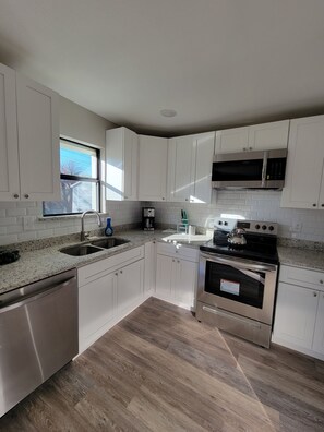 A modern kitchen with white cabinets, a stainless steel dishwasher, a double sink, a coffee maker, a kettle, and a stove. - Spacious 4BE/2BA house with big yard in Downtown Plano (Plano)