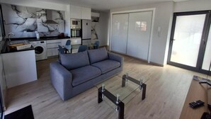 Un salon moderne avec une table basse en verre, un canapé gris et une cuisine en arrière-plan.